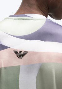 Άτομο που φοράει πολύχρωμο πουκάμισο με αφηρημένα σχήματα και μικρό λογότυπο μαύρου αετού στο πάνω μέρος της πλάτης κοντά στον γιακά.