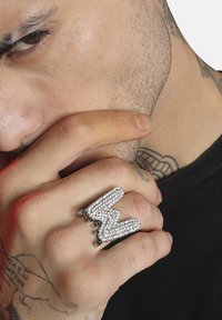 Anillo de plata en forma de "M" estilizada, adornado con strass transparentes y acentos verdes, presentado en una mano con tatuajes visibles.