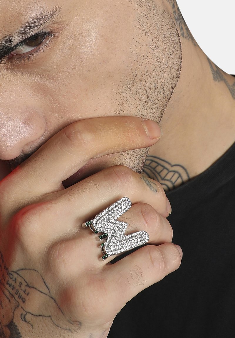 Anillo de plata en forma de "M" estilizada, adornado con strass transparentes y acentos verdes, presentado en una mano con tatuajes visibles.