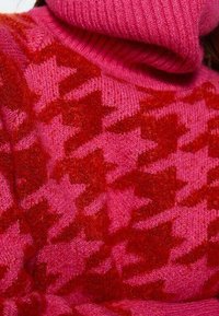 Strickpullover mit hohem Kragen, in leuchtendem Pink mit einem roten Hahnentritt-Muster. Weiche Textur mit gerippten Bündchen und Kragen.
