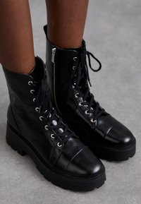 Botas de tobillo de cuero negro con cordones en la parte delantera, cierre de cremallera, puntera redonda y suela gruesa tipo plataforma con textura en la suela.