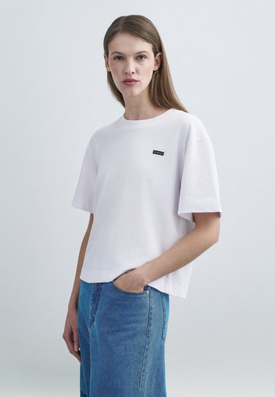 Λευκό T-shirt με κοντά μανίκια και στρογγυλή λαιμόκοψη, με μικρό μαύρο λογότυπο στο στήθος. Ελαφριά υφή υφάσματος. Συνδυάζεται με μπλε τζιν παντελόνι.