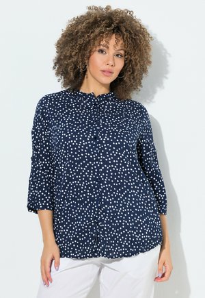 Kvinde med krøllet hår iført en mørkeblå bluse med polkaprikker og hvide bukser, stående mod en hvid baggrund.