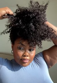 Krøllete, volumøst svart hår stylet opp. En person har på seg en tettsittende lyseblå topp med korte ermer, som fremhever teksturert hud og en ørering.