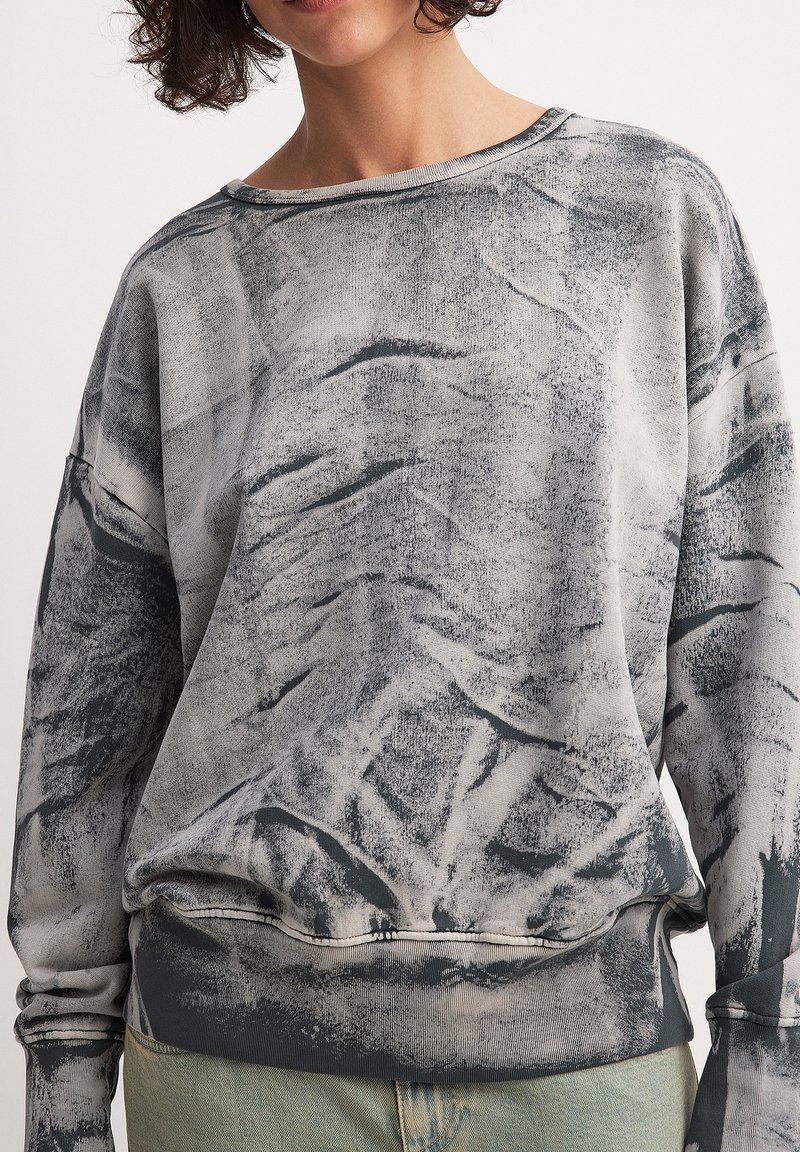 Femme portant un sweat-shirt à manches longues gris et noir marbré avec ourlet élastique et un jean bleu clair sur un fond uni.