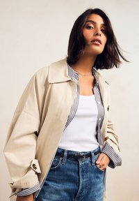 Beež värvi oversize-tüüpi trençkot suure kraega, all triibuline särk ja kõrge vöökohaga sinised teksapüksid. Pronksist nupud ja must vöö detail.