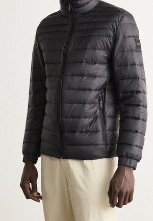 Mann trägt eine schwarze gesteppte Jacke mit Reißverschlusstaschen und elastischen Bündchen, kombiniert mit hellbeigen Hosen, vor einem einfarbigen Hintergrund.