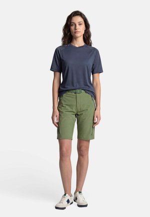 Vrouw staand, gekleed in een donkergrijs kortemouwen T-shirt, groene knielange shorts met ritszakken en witte casual sneakers.