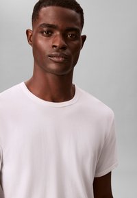 Λευκό βαμβακερό μπλουζάκι με στρογγυλή λαιμόκοψη και κοντά μανίκια, λεία υφή, εφαρμοστή σιλουέτα και διακριτικές ραφές.