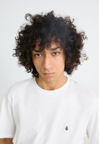 White short-sleeve t-shirt featuring a small black logo on the left chest, worn by a person with curly black hair and a nostril piercing.