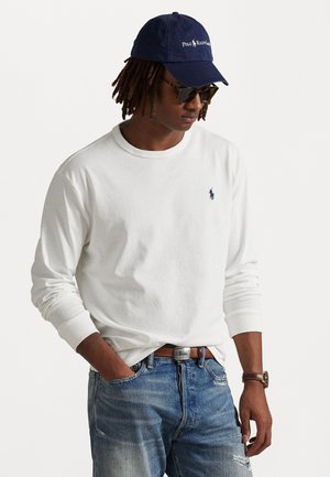 Camisa de algodão branca de manga longa com decote redondo, combinada com jeans claros de lavagem desgastada. Boné azul-marinho com logótipo e óculos de sol escuros.