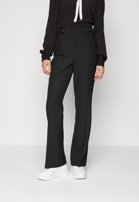 Svarta byxor med rak bendesign, detaljer med veck och sidofickor, tillsammans med en svart långärmad topp och vita sneakers.