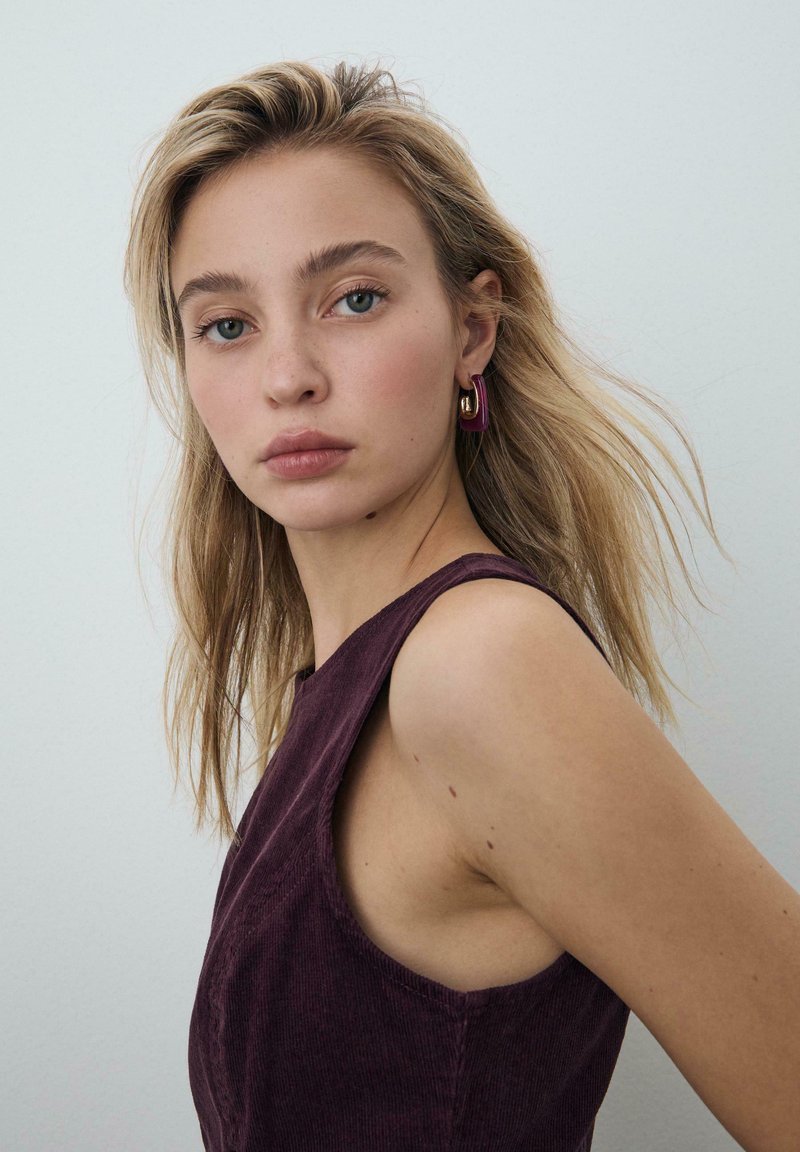 Vinrød ærmeløs top med en struktureret finish og en rundet halsudskæring. Øreringe med øreringe og lilla accenter samt guldfarvet beslag.