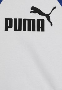 Balta sportinė marškinė su mėlynomis detalėmis, su dideliu juodu "PUMA" logotipu ir juoda puma siluetu. Tekstūruota medžiaga su lygia apdaila.