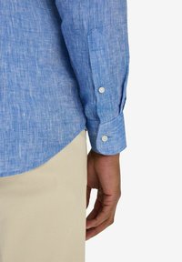 Ljusblå linneskjorta med en subtil textur. Lång ärm, knäppt manschett, böjd nederkant. Hand synlig, bärande beige byxor.