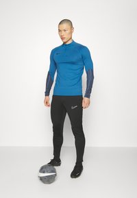 Maglietta sportiva a maniche lunghe blu con maniche stampate nere, abbinata a pantaloni aderenti neri con logo bianco. Scarpe da calcio nere e un pallone da calcio.