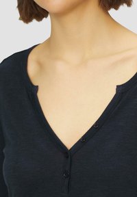 Navyblaues V-Ausschnitt-Oberteil aus strukturiertem Stoff mit einer Knopfleiste. Der Ausschnitt hat eine sanfte Rundung und weiche Konturen.