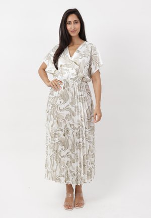 Donna con lunghi capelli scuri che indossa un vestito maxi bianco con motivi floreali beige e tacchi con cinturini trasparenti, in piedi con una mano sui fianchi.