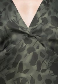 Green floral-patterned fabric with a v-neckline and twisted knot detail. Soft texture with layers; dominant hues are dark green and black.