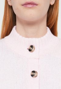Rozā adīts džemperis ar ribotu apkakli, kaklā divas spīdīgas apaļas pogas. Redzama mīksta tekstūra un viegls materiāls.