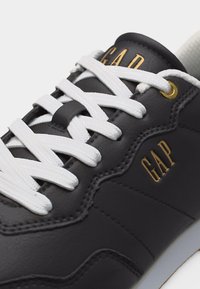 Baskets en cuir noir avec lacets blancs, œillet doré et logo "GAP" en or sur la languette et le côté.