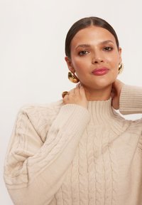 Beżowy sweter z dzianiny o wysokim kołnierzu, z prążkowanymi mankietami i luźnym krojem, zestawiony z dużymi złotymi kolczykami w kształcie kół oraz złotym pierścionkiem.