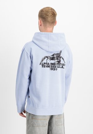 Blonder Mann trägt eine hellblaue Kapuzenjacke mit einem schwarzen Skorpion und dem Text "Alpha Industries Tennessee U.S.A. 1959" auf dem Rücken.
