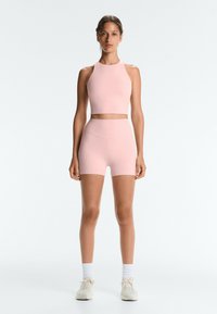 Woman standing facing forward wearing light pink athletic crop top and matching shorts with white socks and sneakers on plain background.