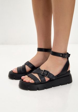 Black leather sandals with multiple wide straps, adjustable ankle buckle, and textured chunky sole. Open-toe design shows toenails.