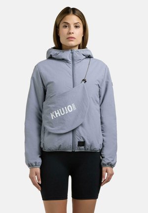 Kvinde iført en lysegrå hættetrøje og sorte cykelshorts, bærende på en matchende crossbody taske med teksten "KHUJO 2001".