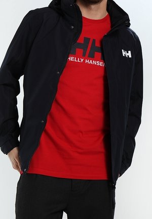 Homme portant une veste noire zippée avec un logo blanc sur un t-shirt rouge Helly Hansen et un pantalon noir, debout devant un fond uni.