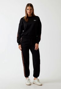 Hanorac negru din bumbac cu manșete striate și logo brodat. Pantaloni negri asortați, cu dungi laterale maro și tiv elastic la gleznă.