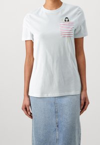 Kvinna klädd i en vit T-shirt med en rödrandig handduk och en liten kvinnografik på bröstet, kombinerad med en ljusblå denimkjol.