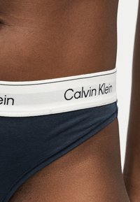 Gros plan sur la hanche d'une personne portant des sous-vêtements noirs Calvin Klein avec une ceinture blanche et le nom de la marque visible.