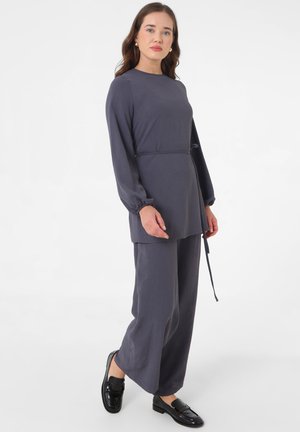 Vrouw draagt een donkergrijze tuniek met lange mouwen en een taillekoord, gecombineerd met bijpassende wijde broekspijpen en zwarte loafers.