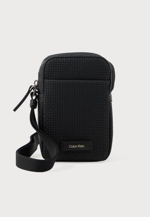 Bolsa de crossbody tejida negra con bolsillo frontal, cierre de cremallera, correa para la muñeca y placa con el logo de Calvin Klein en la parte frontal.