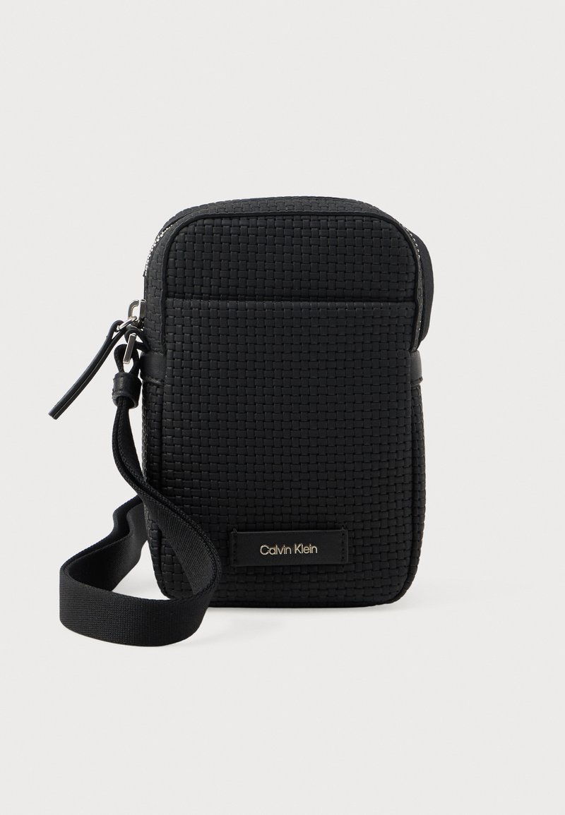 Bolsa de crossbody tejida negra con bolsillo frontal, cierre de cremallera, correa para la muñeca y placa con el logo de Calvin Klein en la parte frontal.