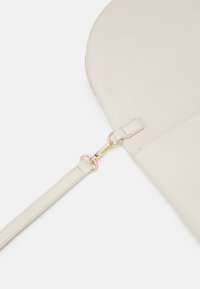 Beige leren schoudertas met een textuur oppervlak, een sluiting van goudkleurig metaal en een afneembare band. Heeft een afgerond, minimalistisch ontwerp.