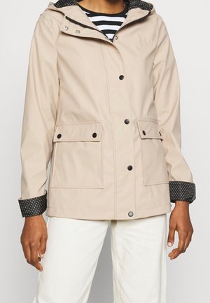 Veste à capuche beige avec boutons-pression noirs et poignets intérieurs à pois, portée sur un haut rayé noir et blanc et un pantalon blanc.