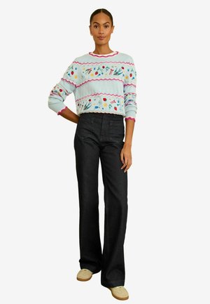 Vrouw draagt een lichtblauwe trui met kleurrijke bloemrijke borduurwerk en roze bies, gecombineerd met zwarte, hoogtallige, wijde broekspijpen en beige schoenen.