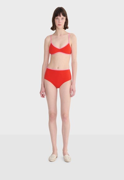 Femme debout, portant un bikini rouge avec des bords roses et des chaussures beige à enfiler, sur un fond gris clair uni.