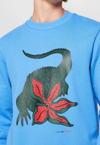 Persona que lleva un sudadera azul con un gráfico de cocodrilo verde combinado con una flor roja en el pecho, que presenta los logotipos de Lacoste y Netflix.