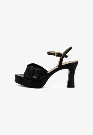 Sandaletto nero con plateau e tacco alto, con cinturino anteriore annodato, cinturino alla caviglia e fibbia dorata su sfondo bianco.
