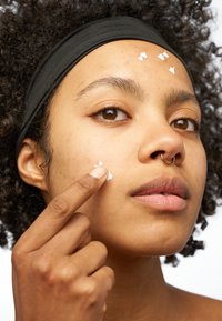 Produit de soin de la peau appliqué sur un visage, présentant un teint brun, des cheveux bouclés et un bandeau noir. Texture crémeuse blanche visible sur la peau.