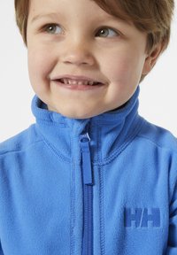 Modrý flísový svetr s vysokým límcem, se zapínáním na přední zip a texturovaným povrchem. Na hrudi má tmavě modré logo.