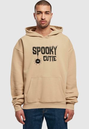 Beige hættebluse med lomme foran og sort "Spooky Cutie" grafik, der viser en tegneserieedderkop. Blødt bomuldsblandingsstof.