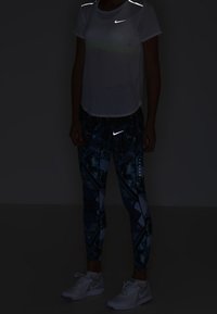 Maglietta sportiva riflettente Nike e leggings a fantasia in colori scuri, indossati con sneakers bianche. Dettagli riflettenti sulla maglietta e sui leggings.