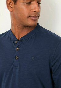 Marineblaue Henley-Bluse mit rundem Ausschnitt, einer Drei-Knopf-Leiste und subtiler Textur; enthält ein kleines, gesticktes Logo auf der Brust.