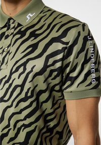 Olivgrön polo med ett mönster av svarta tigerränder. Har en krage och sidotryck av märkesnamnet i vitt på ärmen.