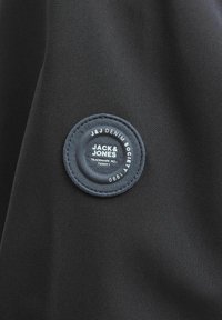 Zwart stof met een cirkelvormige blauwe patch; tekst leest "JACK & JONES" en "J & J DENIM SOCIETY," met verhoogde textuur en stiksels.